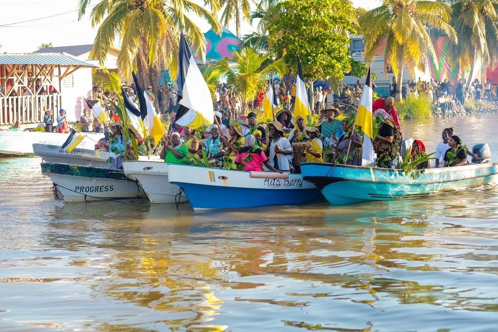 Boats of people in traditional Garifuna clothing carrying flags on a river.